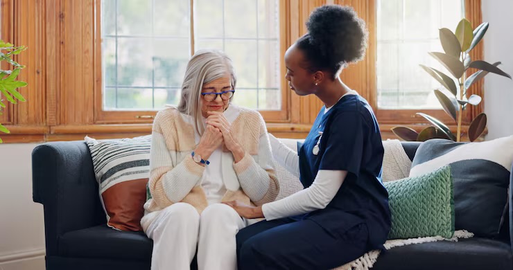 Caregiver sitting beside an older woman on a sofa, offering a reassuring hand