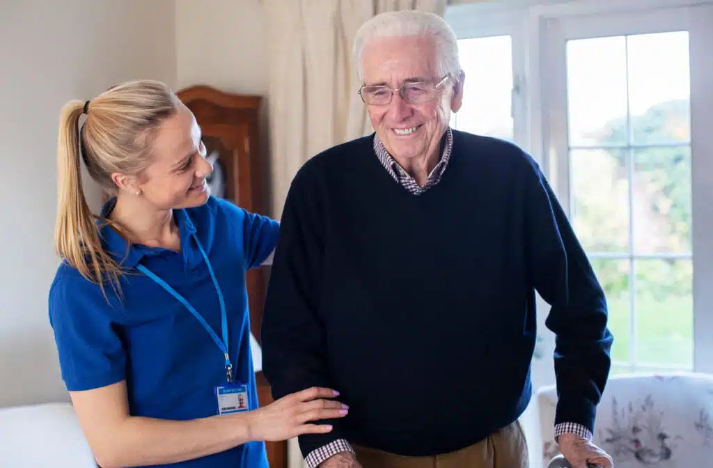 Caregiver in blue steadying an older man as he walks across his living room